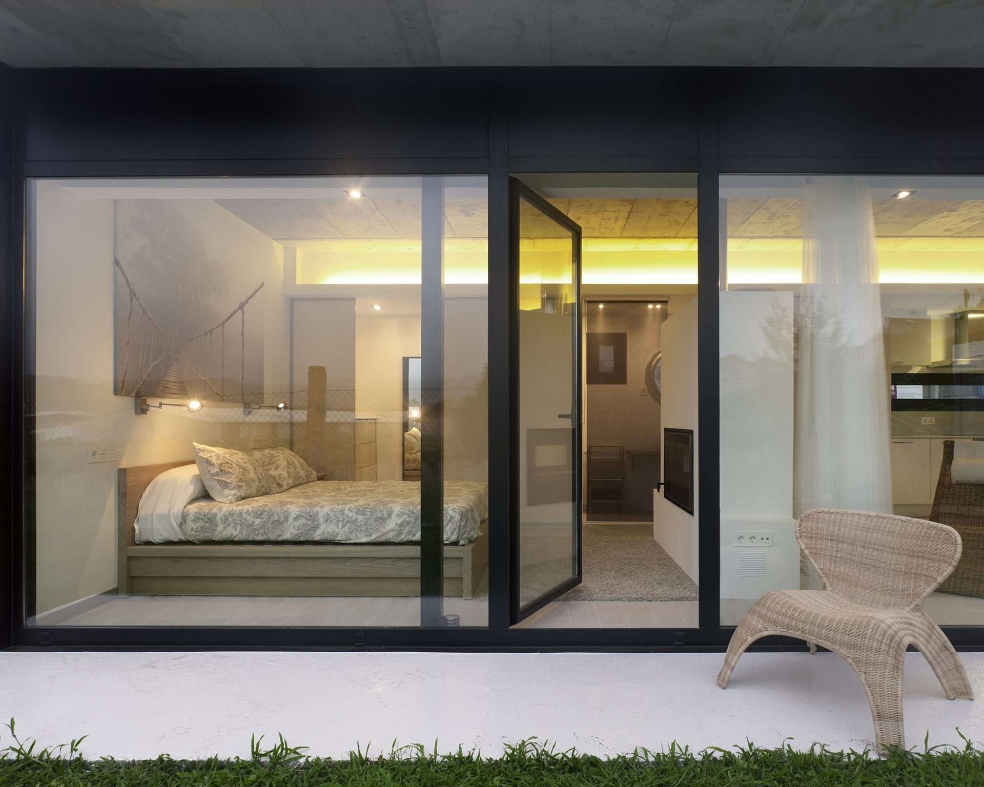 Glass windows line one side of the home and a glass door is open leading to a bedroom.