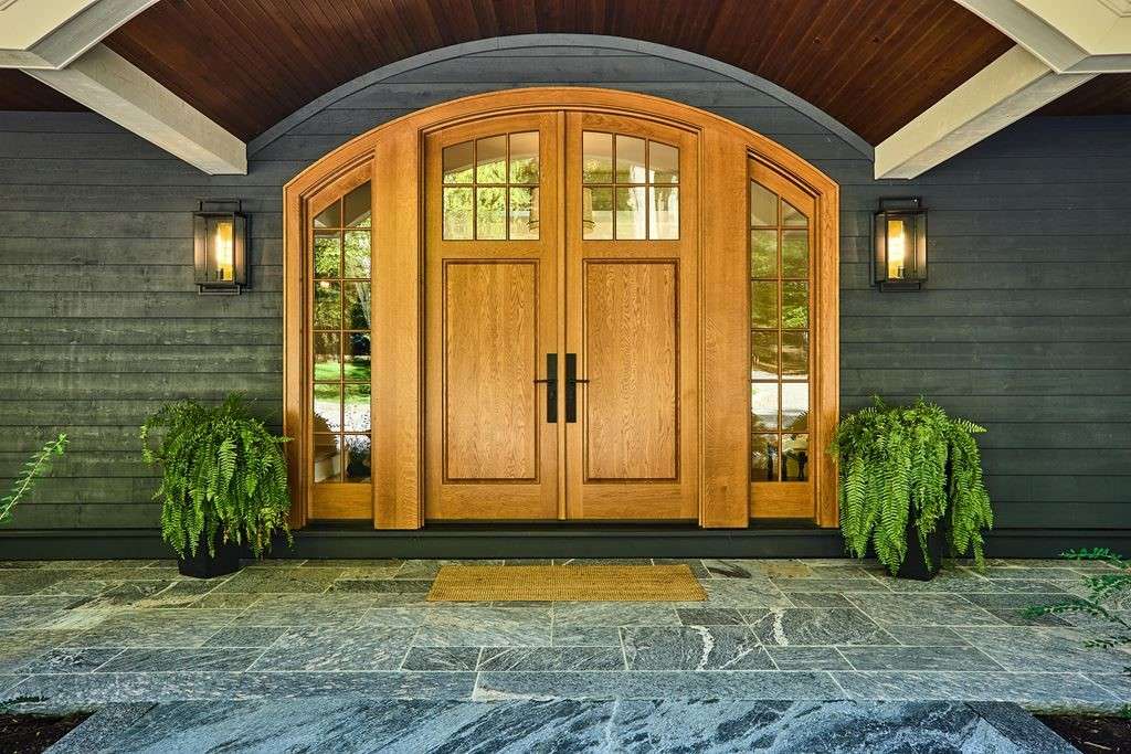 A grand, arched wood entry door with decorative sidelites and transom, framed by dark vertical siding and flanked by large potted ferns on a stone porch.