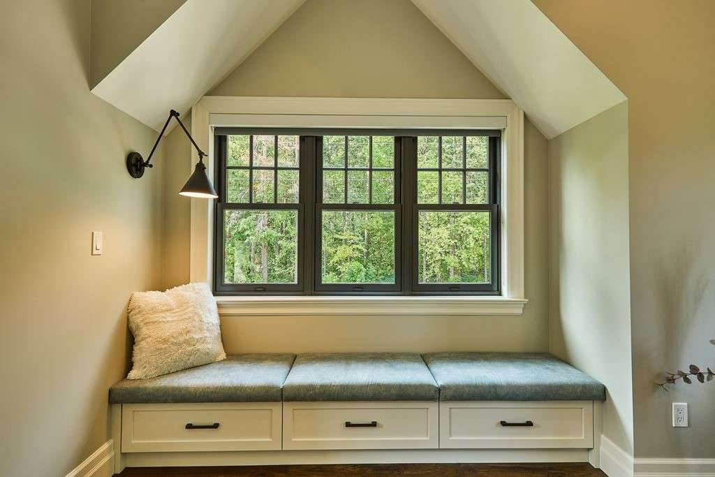 A cozy window seat nook with a black-framed grid window overlooking a green wooded area, built-in drawers beneath the bench, and a wall-mounted lamp.