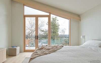 A bright, modern bedroom with a sloped ceiling features large wood-framed glass doors and windows opening to a balcony with outdoor seating.