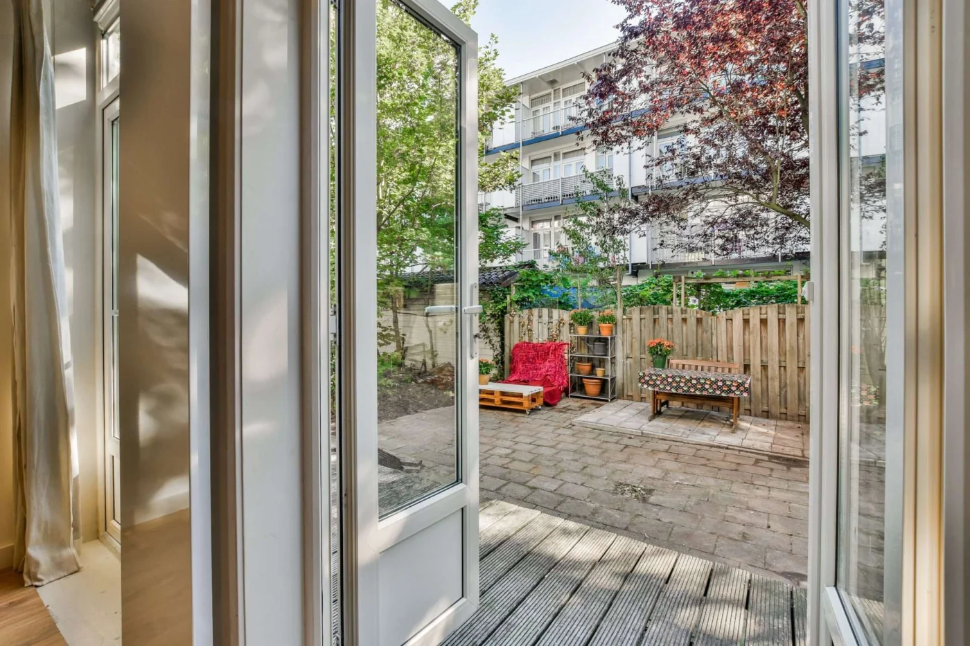 Impeccable Patio Doors | Cold Springs, NY | Rockman Windows and Doors Open white patio doors leading to a fenced courtyard with paver patio, bench, potted plants, leafy trees, wood fence, and apartments beyond.