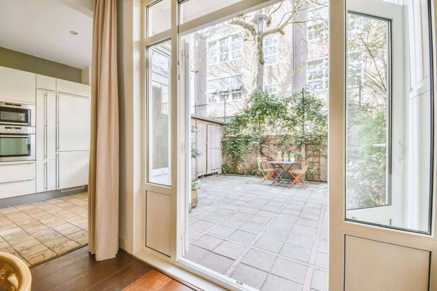 Terrific Patio Doors | Cold Springs, NY | Rockman Windows and Doors White patio doors open from a bright kitchen to a stone courtyard with red chairs, bistro table, ivy wall, tall tree, and brick building.